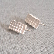 A pair of textured silver stud earrings with a grid-like pattern, resting on a neutral gray linen fabric background.