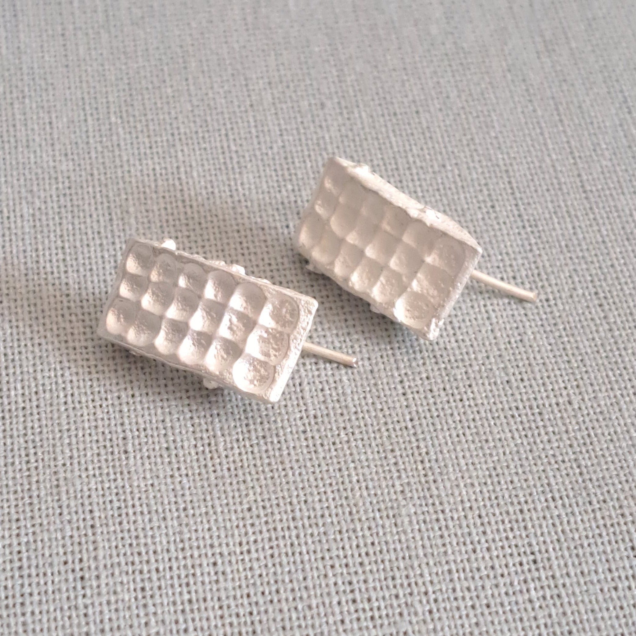 A pair of textured silver stud earrings with a grid-like pattern, resting on a neutral gray linen fabric background.