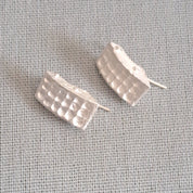 A pair of textured silver post earrings with a wavy, segmented design, displayed on a light gray linen fabric background.