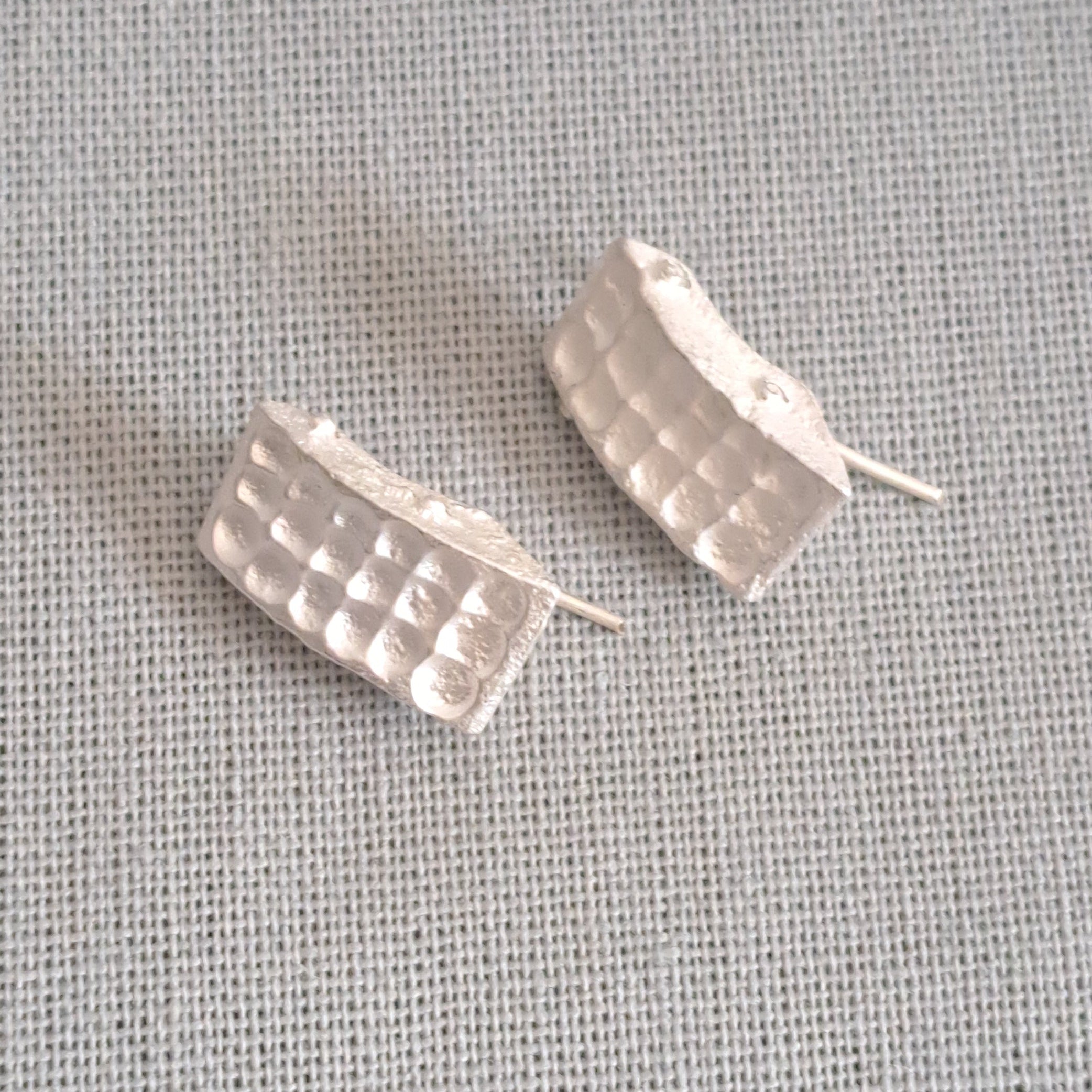 A pair of textured silver post earrings with a wavy, segmented design, displayed on a light gray linen fabric background.