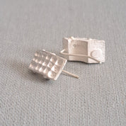 A pair of small silver stud earrings with a textured, grid-like top design, resting on a light gray linen fabric background.