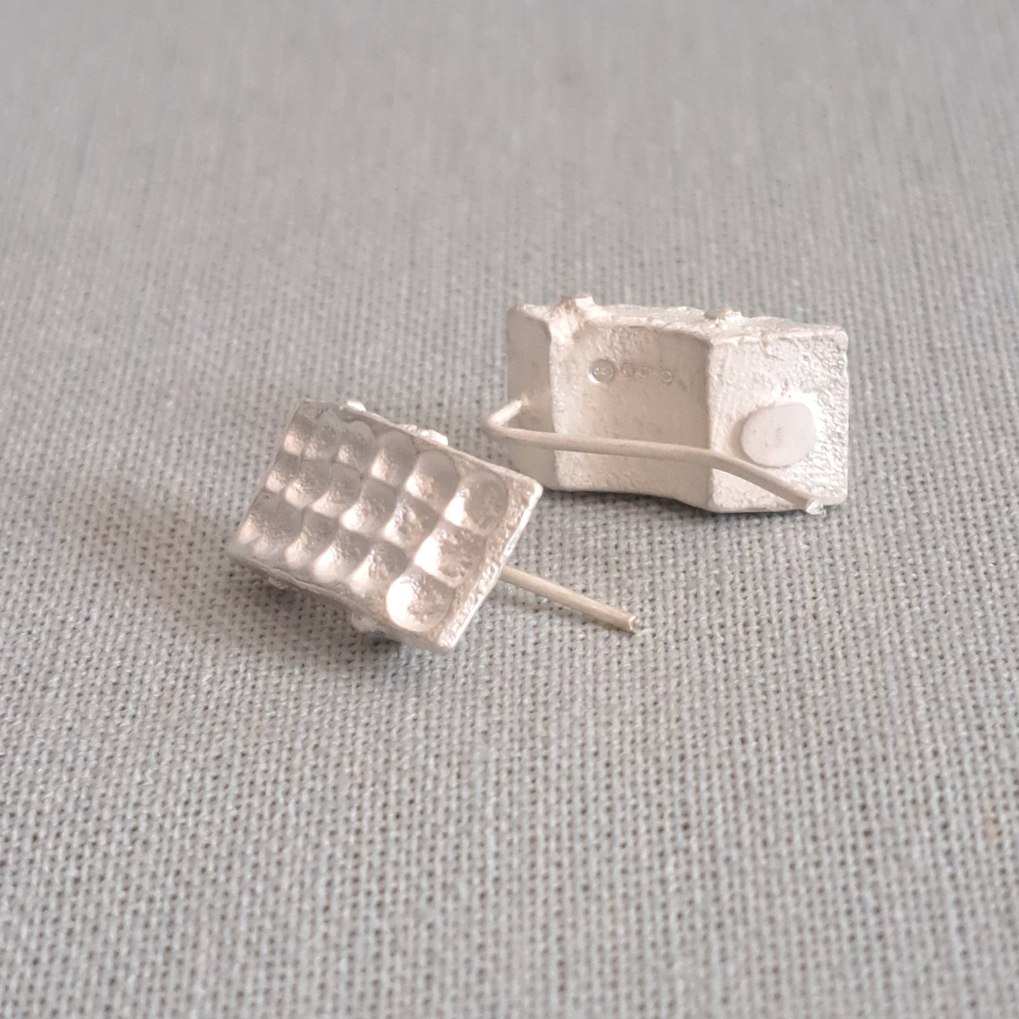 A pair of small silver stud earrings with a textured, grid-like top design, resting on a light gray linen fabric background.
