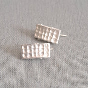 A pair of small silver stud earrings with a textured, grid-like rectangular design, displayed on a neutral gray fabric background.