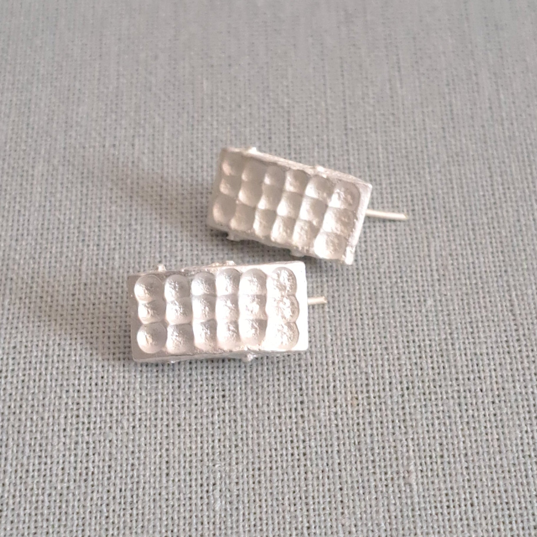 A pair of small silver stud earrings with a textured, grid-like rectangular design, displayed on a neutral gray fabric background.