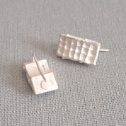 A pair of textured silver stud earrings with a hammered rectangular design, displayed on a neutral gray fabric background.