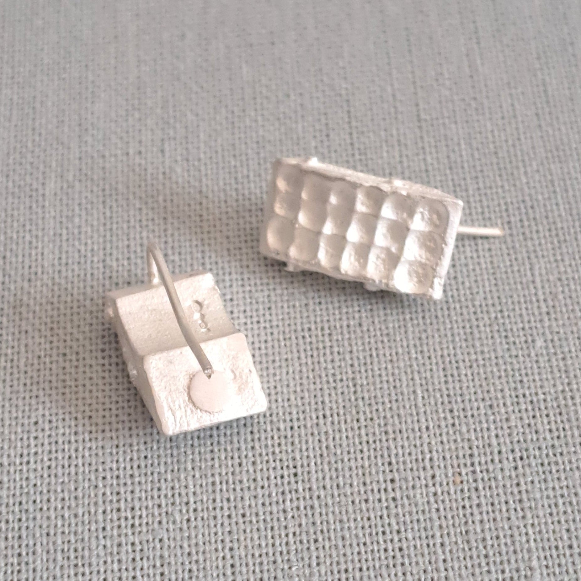 A pair of textured silver stud earrings with a hammered rectangular design, displayed on a neutral gray fabric background.