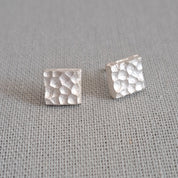 A pair of small square stud earrings with a textured hammered finish, shown on a neutral gray textured fabric background.