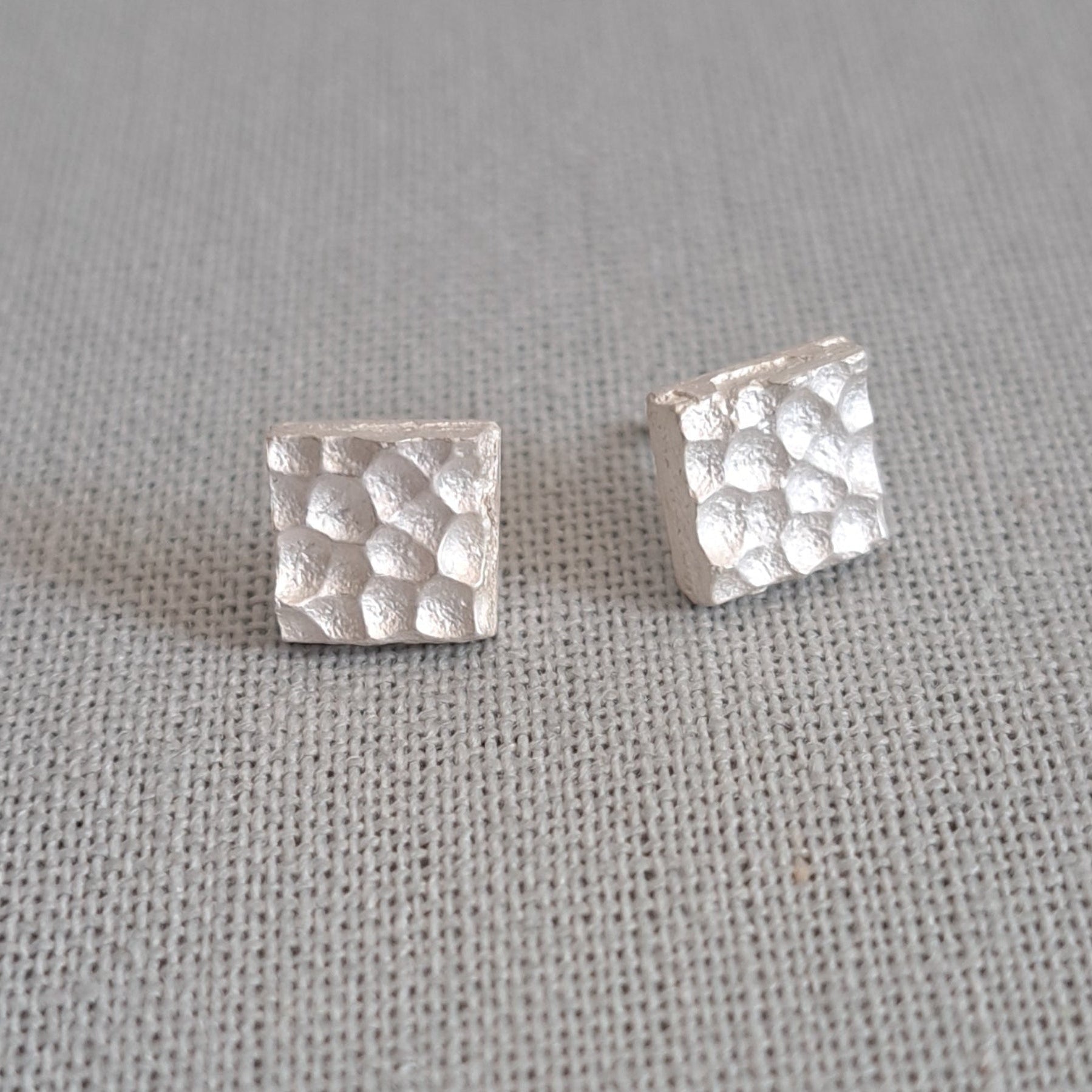 A pair of small square stud earrings with a textured hammered finish, shown on a neutral gray textured fabric background.