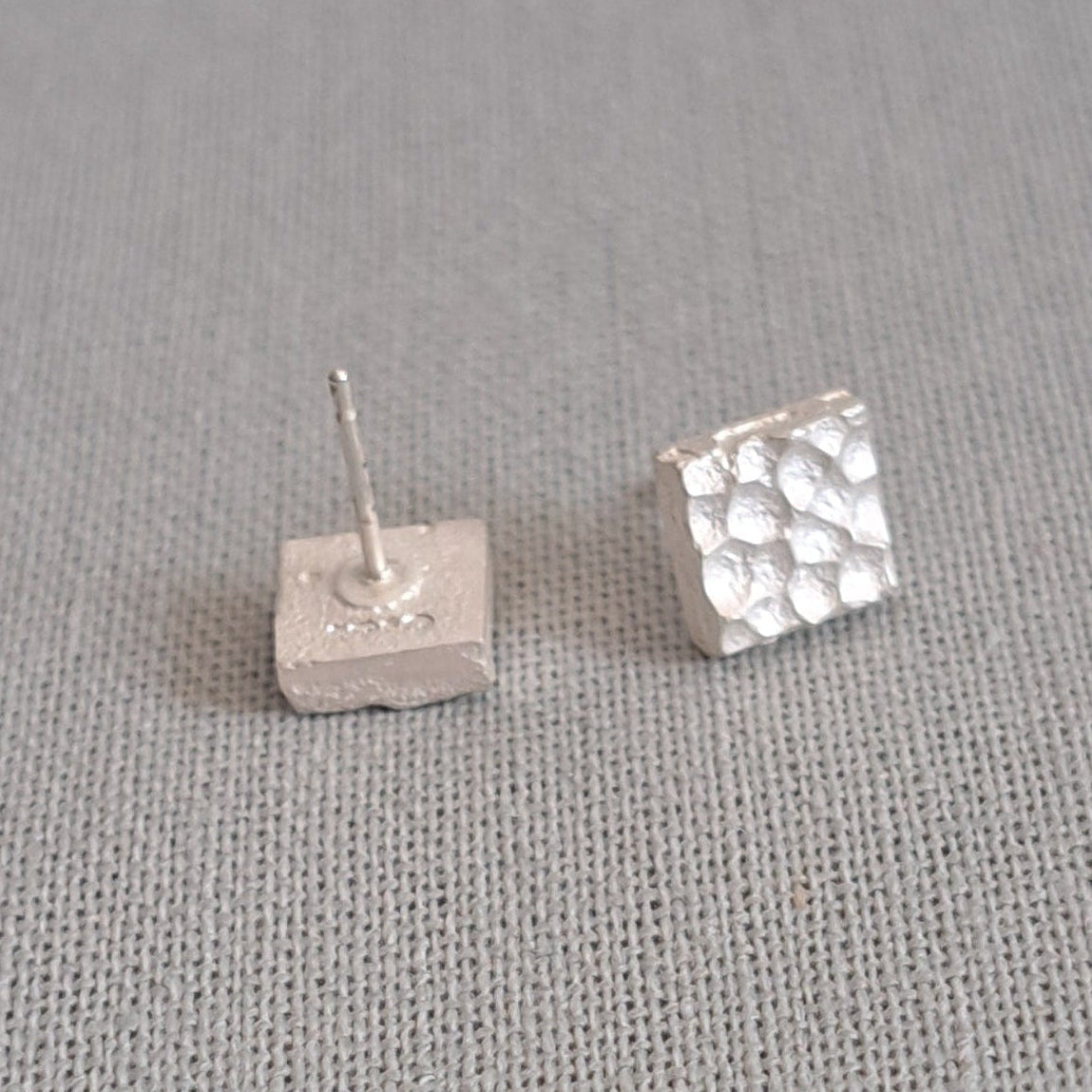 A pair of small silver stud earrings on a textured gray fabric background. One earring is plain, and the other has a textured, dimpled surface.