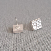 A pair of small silver stud earrings on a textured gray fabric background. One earring is plain, and the other has a textured, dimpled surface.