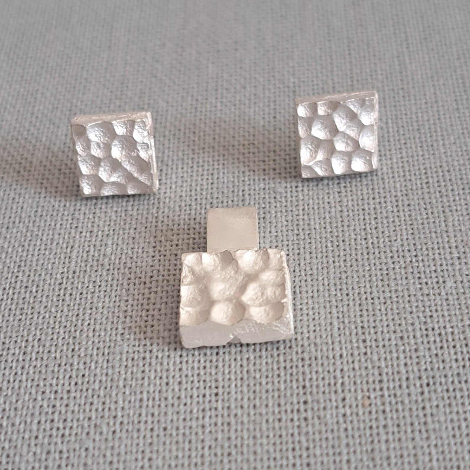 A pair of silver stud earrings with a hammered square design, displayed on a textured gray fabric background.