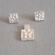 A pair of silver stud earrings with a hammered square design, displayed on a textured gray fabric background.