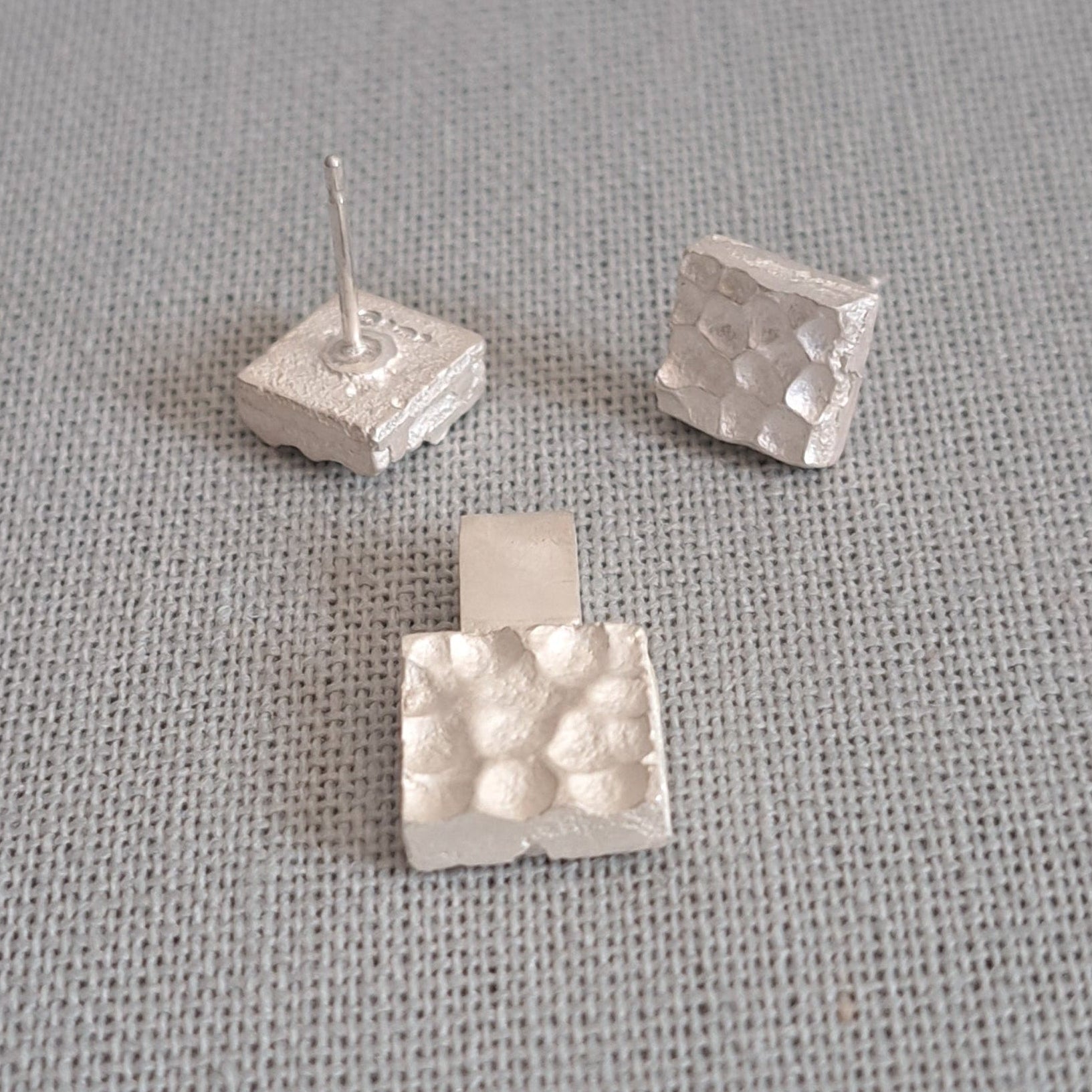 Three small silver textured cube stud earrings on a gray fabric background.