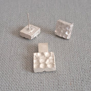 Three small silver textured cube stud earrings on a gray fabric background.