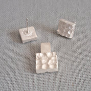 Three small square silver stud earrings with a textured, dimpled surface, displayed on a gray woven fabric background.