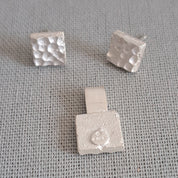 Three small silver jewelry pieces with textured square designs, including two stud earrings and one pendant, displayed on a gray textured fabric background.