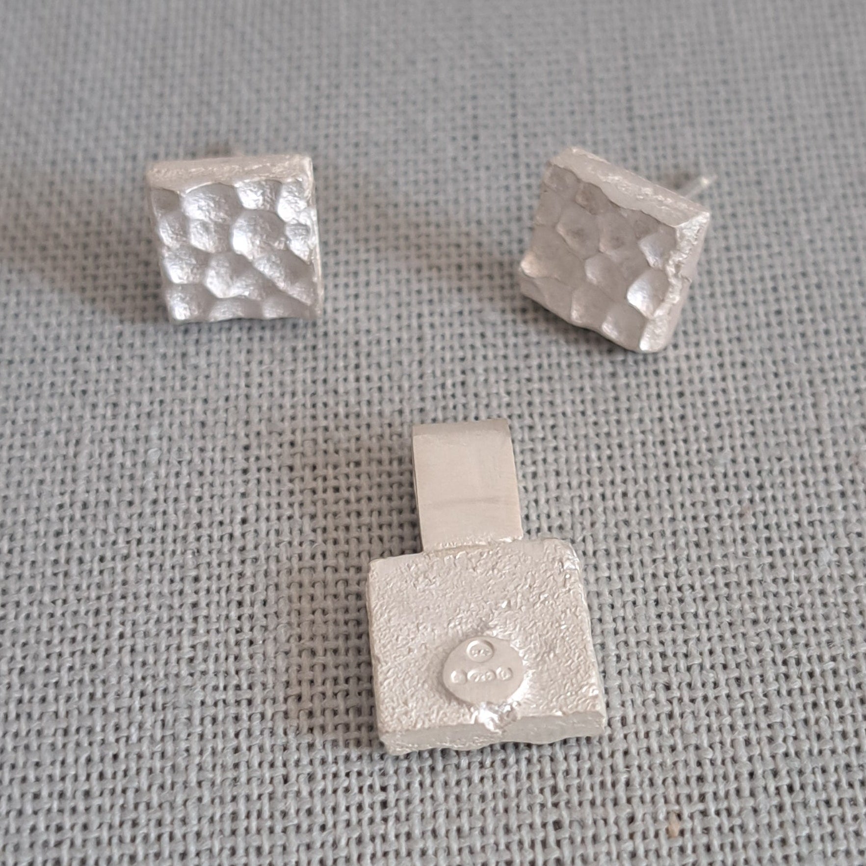 Three small silver jewelry pieces with textured square designs, including two stud earrings and one pendant, displayed on a gray textured fabric background.