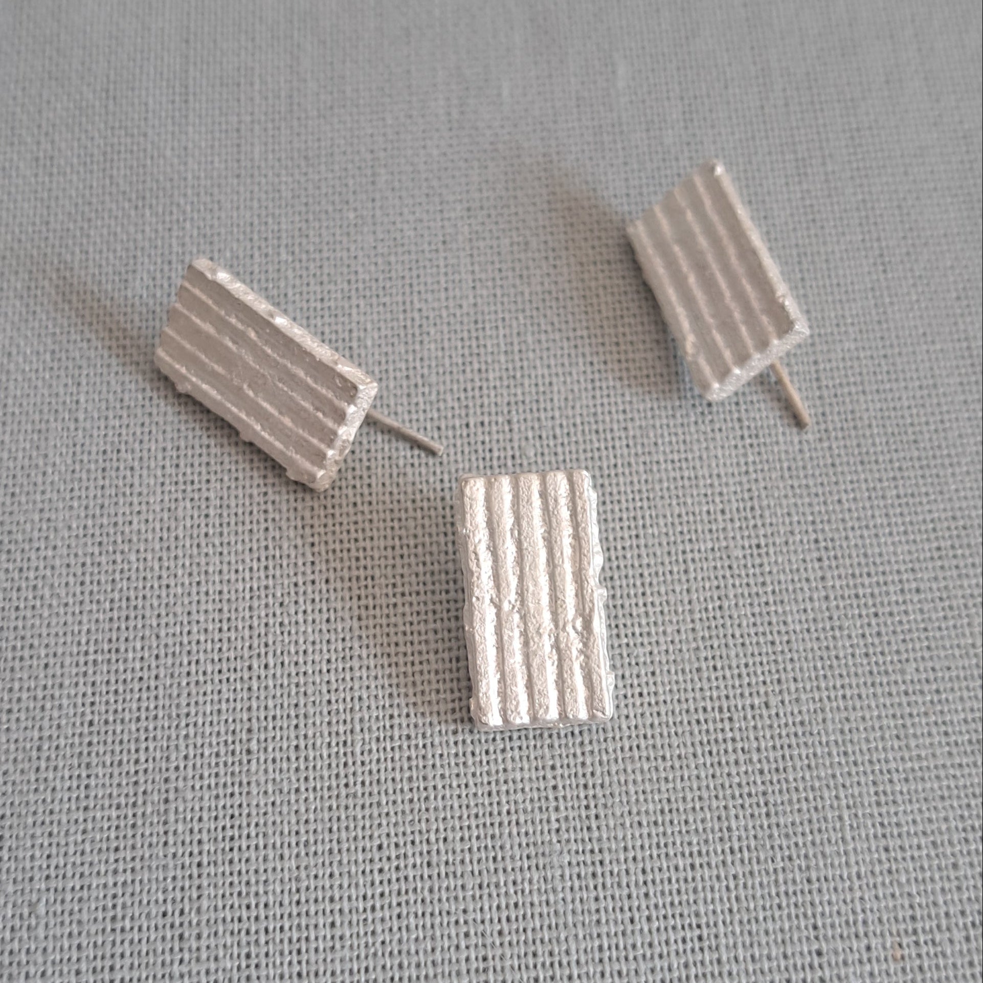 Three small rectangular stud earrings with vertical ridges, shown on a textured light gray fabric background.