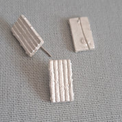 A pair of silver-toned rectangular stud earrings with vertical textured ridges, shown on a textured gray fabric background.