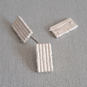 A pair of small rectangular stud earrings with a textured vertical ribbed pattern, shown on a neutral gray textured fabric background.