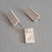 A pair of small rectangular stud earrings with a ribbed textured surface, shown on a gray fabric background. One earring is shown from the front and the other from the side, revealing the post.