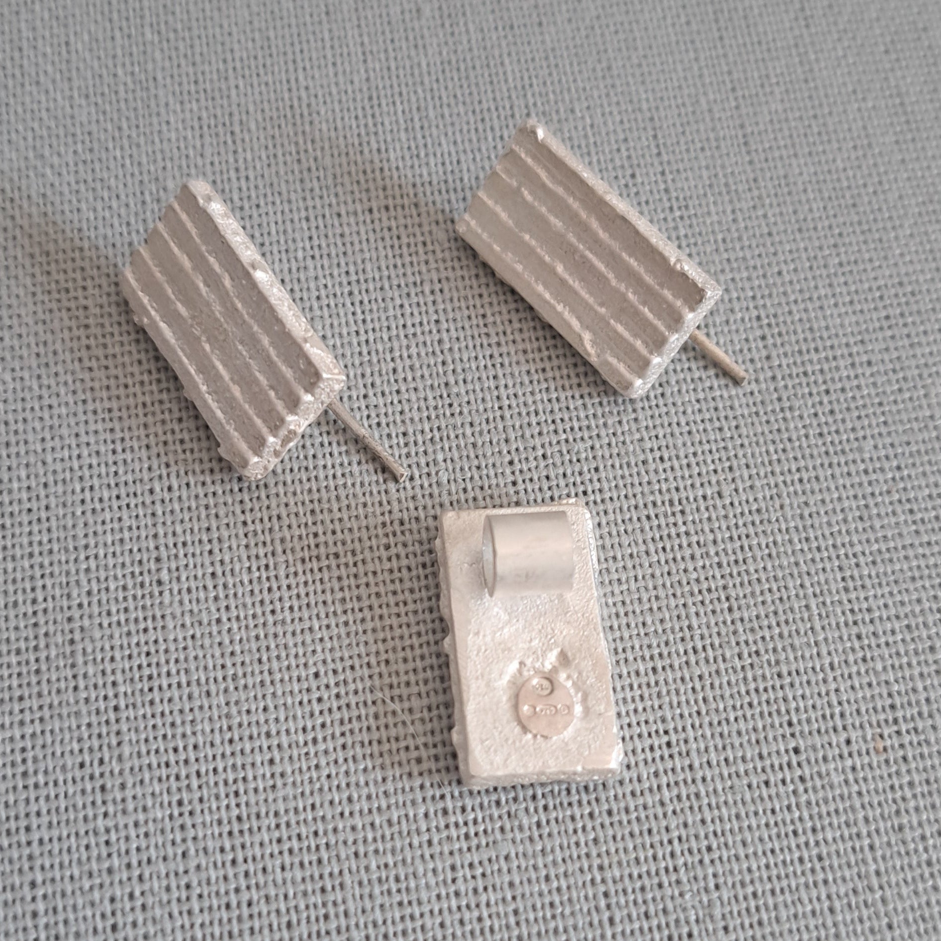 A pair of small rectangular stud earrings with a ribbed textured surface, shown on a gray fabric background. One earring is shown from the front and the other from the side, revealing the post.