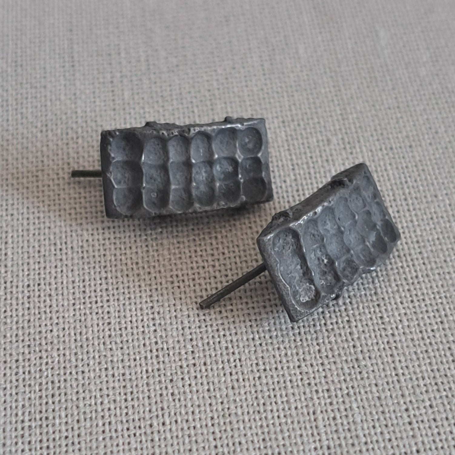 A pair of small rectangular stud earrings with a textured, ribbed metallic surface in a dark silver or gunmetal finish, resting on a light beige linen fabric background.