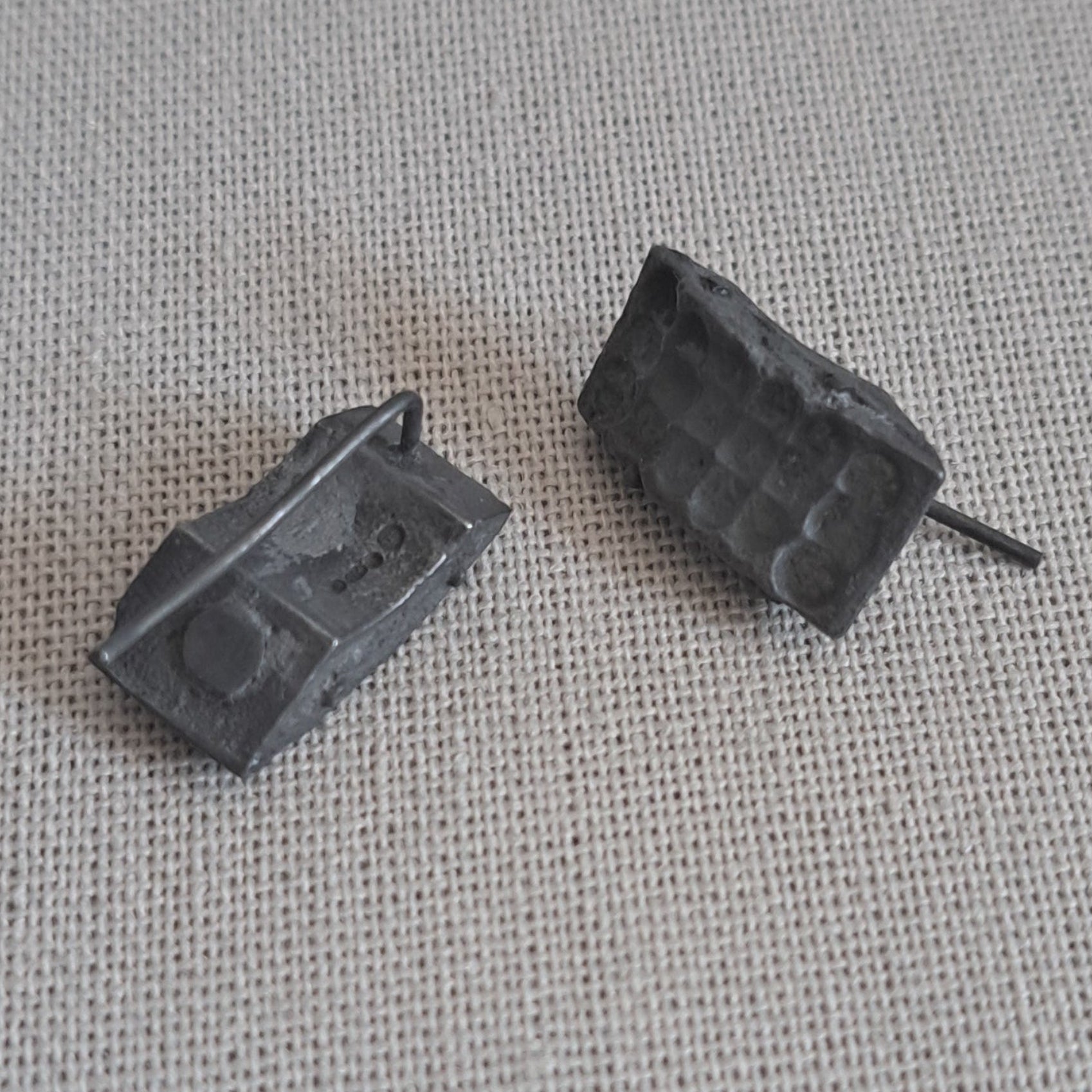 A pair of small dark gray rectangular stud earrings with a textured, blocky design, resting on a light gray woven fabric background.