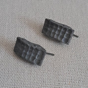 A pair of small black textured stud earrings with a grid-like pattern, resting on a light gray linen fabric background.