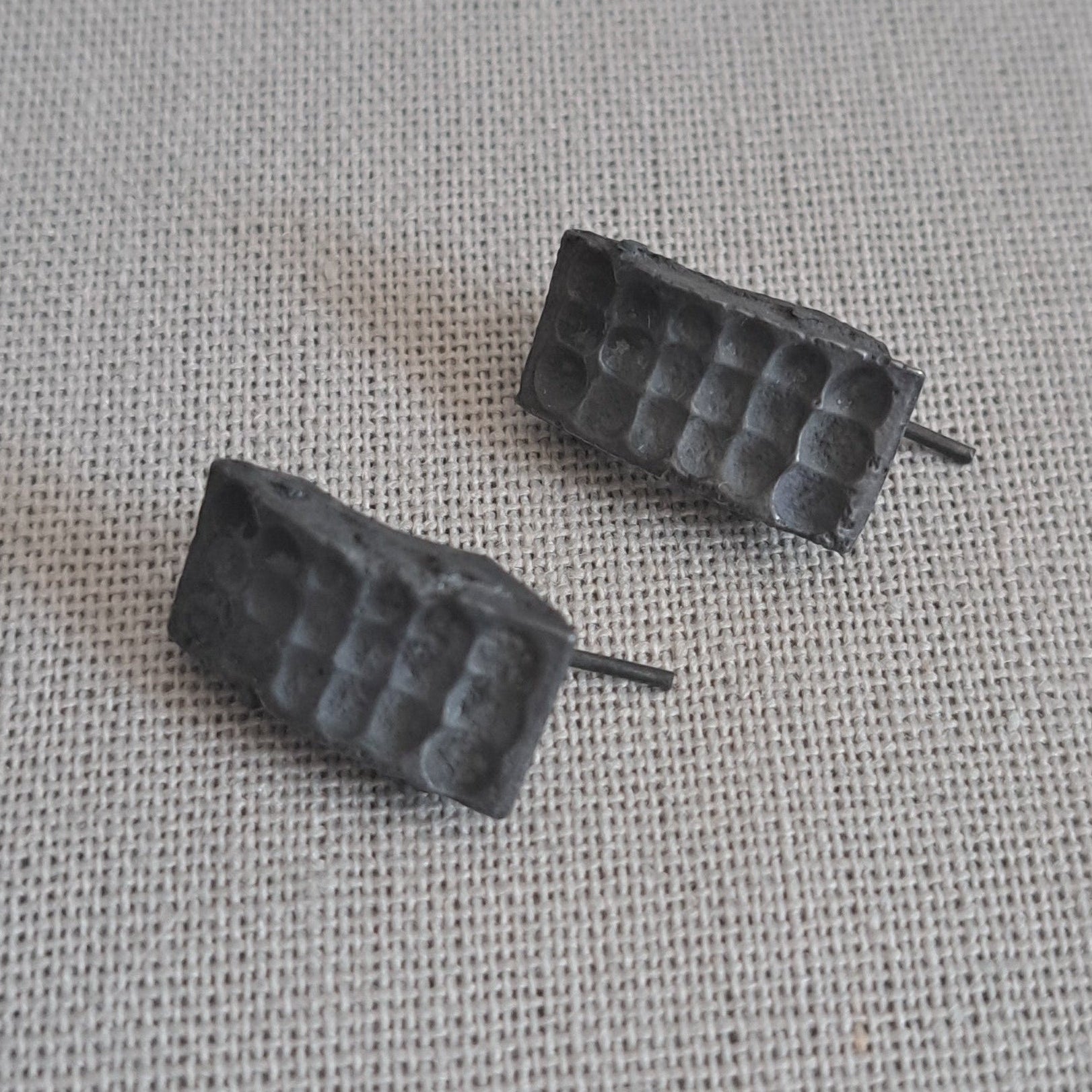 A pair of small black textured stud earrings with a grid-like pattern, resting on a light gray linen fabric background.